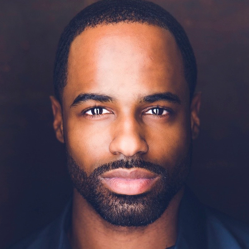 A close-up portrait of a man with a short beard and mustache, closely cropped hair, and brown eyes. He has a serious expression and is wearing a dark shirt. The background is softly blurred in brown tones, drawing focus to his face.