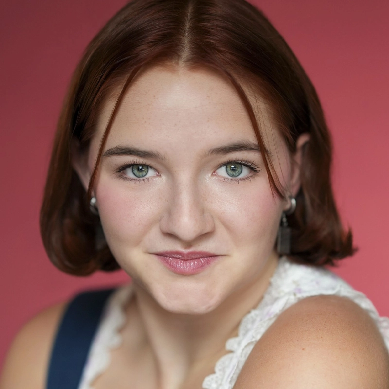 Rebecca Dral, a young woman with short brown hair and green eyes, smiles gently at the camera. She wears a white lace top and small hoop earrings. The solid pink background enhances the warm and inviting feel of the portrait.