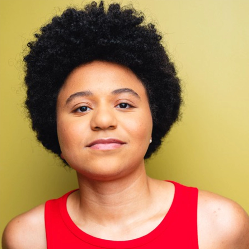 A person with short, curly black hair and light brown skin wears a sleeveless red top. They look directly at the camera with a subtle smile reminiscent of Monster Mike Welch. The background is a solid, muted yellow, lit brightly and evenly.