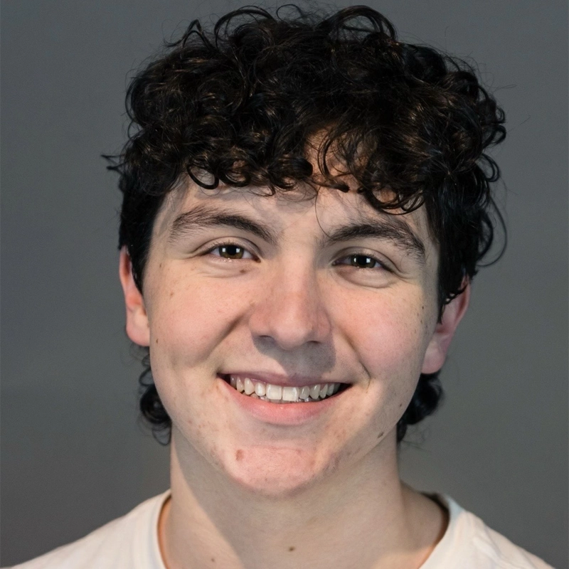 Aydan Eatchel, a young person with short, curly dark hair, smiles at the camera. They have light skin, dark eyes, and are wearing a white shirt. The solid gray background and even lighting highlight their face clearly.