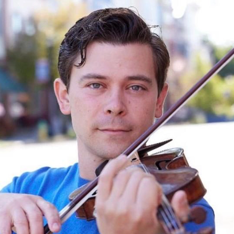 A man with short brown hair, identified as Edmund Bagnell, wearing a blue shirt is playing a violin outdoors. He looks directly at the camera, holding the violin under his chin with one hand and the bow in the other amidst a blurred background.