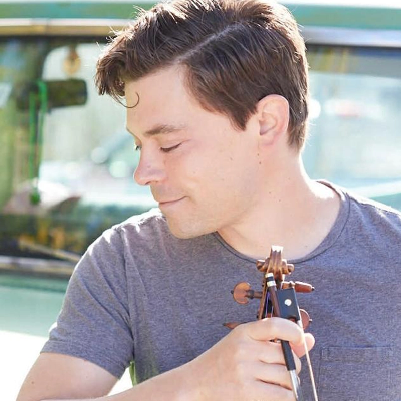Edmund Bagnell, a man with short brown hair and fair skin, wearing a gray t-shirt, holds a violin close to his face and looks down with a gentle expression. Bright sunlight illuminates him, with a blurred green vehicle in the background.