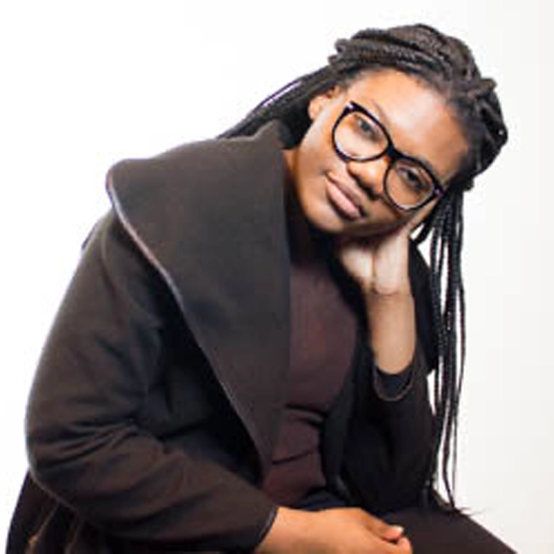 A young woman with long braids and black glasses sits against a white background. Viani Edwards wears a dark coat and rests her head on her hand, leaning slightly forward with a thoughtful expression.