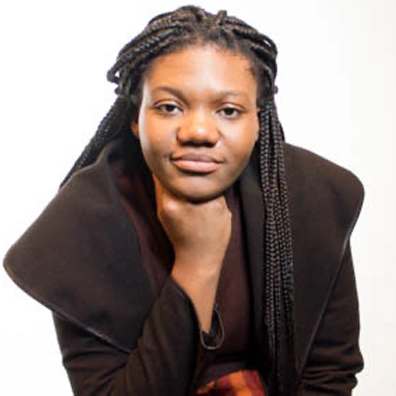 Viani Edwards, with long, braided hair and a dark coat, poses confidently against a plain white background. Resting their chin on one hand, they look directly at the camera with a slight smile. The lighting is bright and even.