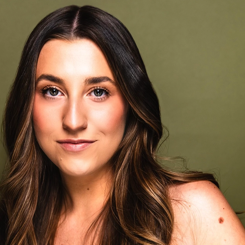 Elena Garcia, a young woman with long, wavy brown hair and blue eyes, poses against a plain olive green background. She has natural makeup, a gentle smile, and her bare shoulder is slightly visible in the foreground.