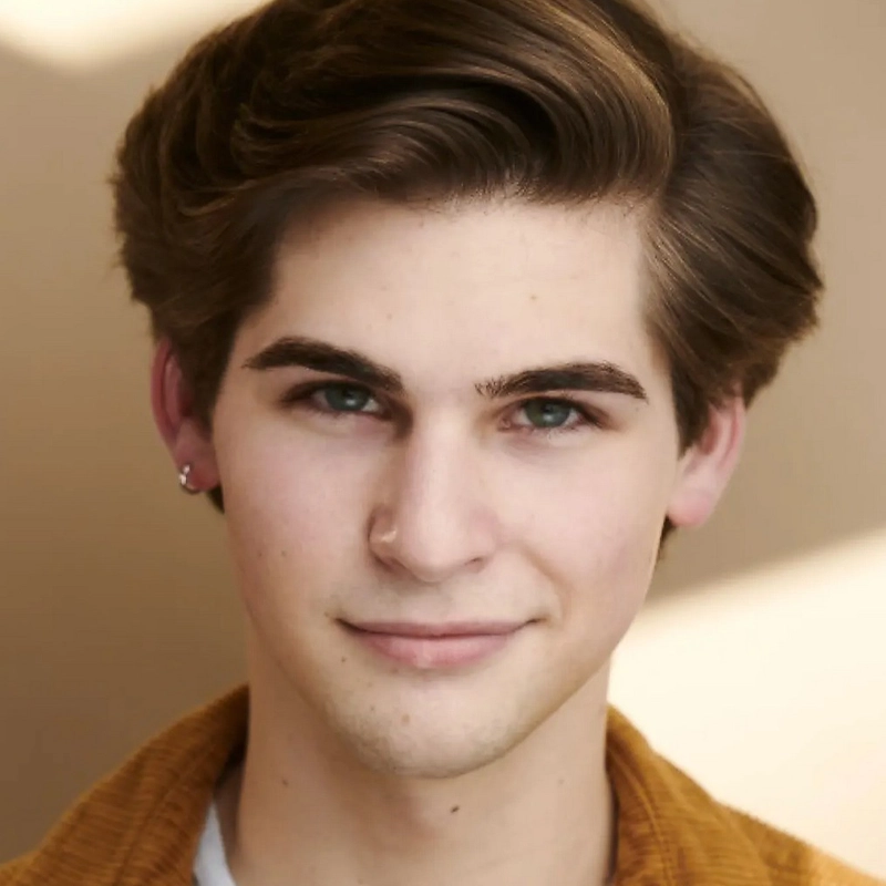 A young person with fair skin, short brown hair styled to the side, green eyes, and a subtle smile looks at the camera. They wear a mustard yellow jacket and a small silver earring in the left ear. The background is softly blurred beige.