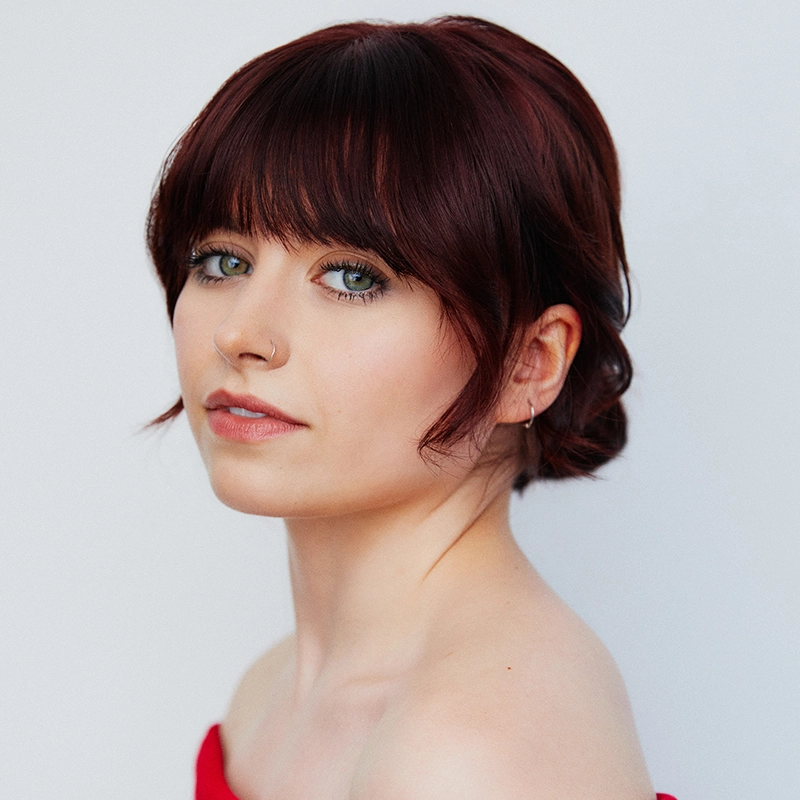 Casey Martin Klein, a young woman with fair skin and short, dark auburn hair styled in loose waves with bangs, has green eyes, subtle makeup, a nose ring, and small hoop earring. She wears an off-shoulder red top against a plain light background.