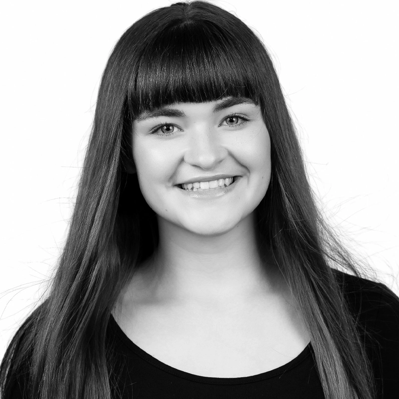 Claire Elliott, a young woman with long straight hair and blunt bangs, smiles at the camera. She is wearing a black top and is photographed against a plain white background in a striking black-and-white portrait.