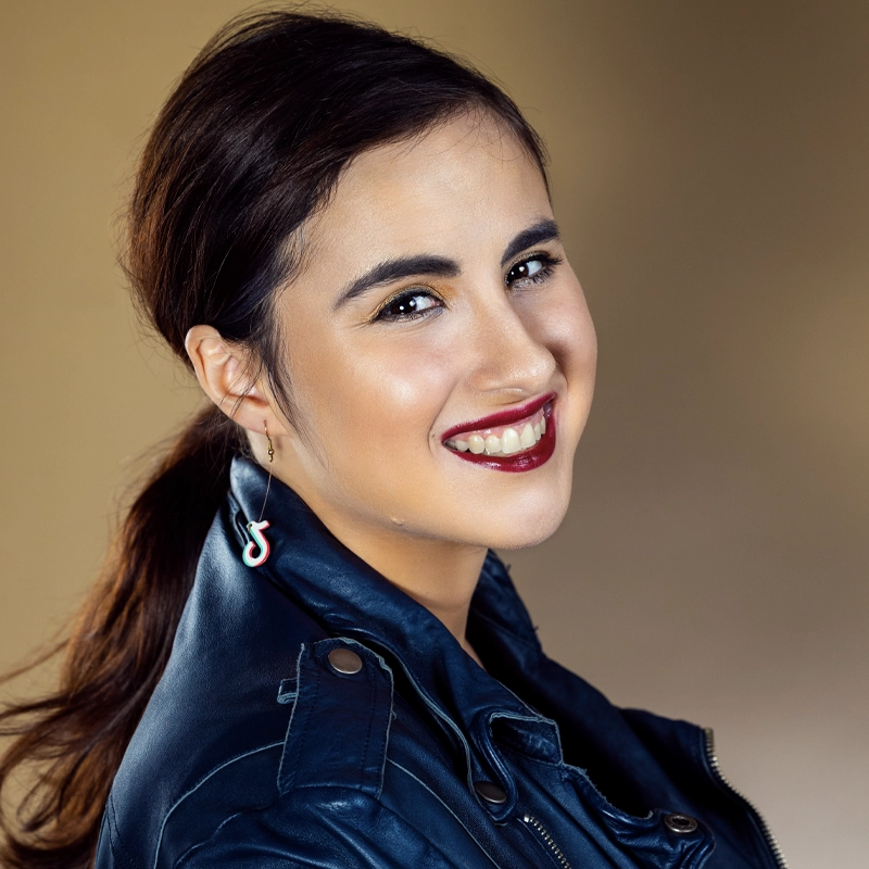 A young woman with dark hair pulled back smiles warmly. She wears bold red lipstick, dark eye makeup, a black leather jacket, and unique safety pin earrings with colored accents—channeling Amy Erlanger style against a soft, warm beige background.