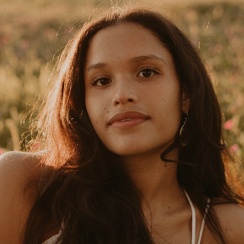 Aubrie Ofner, a young woman with long, dark brown hair and warm brown skin, stands outdoors in soft sunlight. She has a calm expression, wears a white halter top and gold earrings. The blurred background shows green grass and pink wildflowers.
