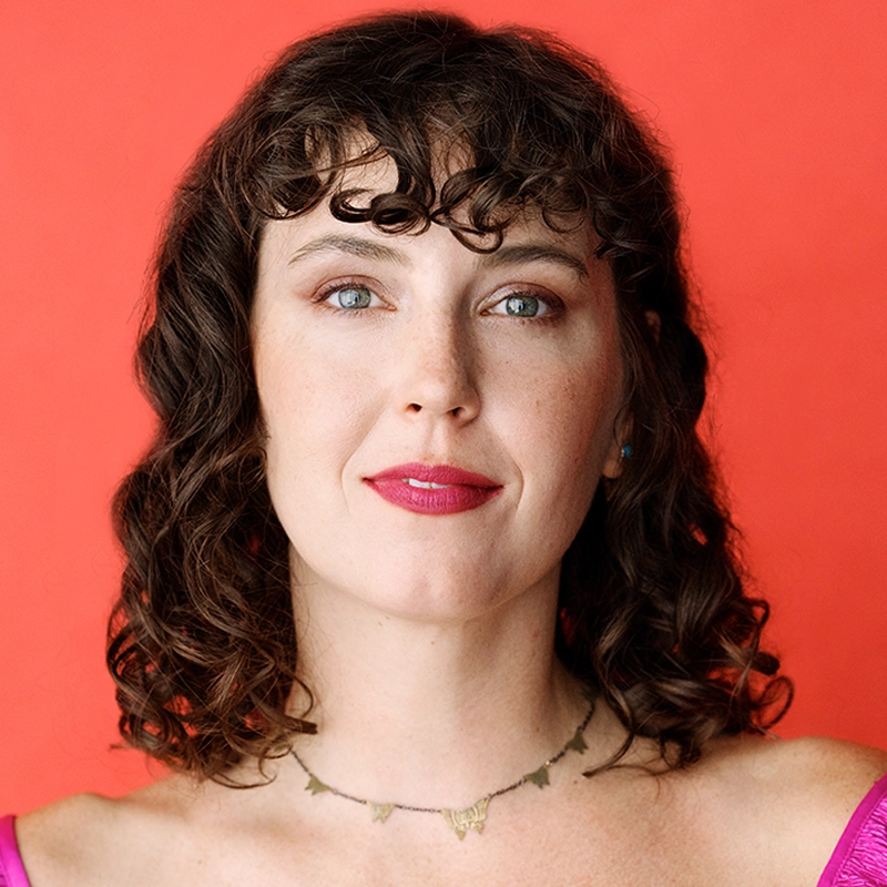 A woman with curly brown hair and blue eyes is wearing a pink top and a delicate necklace. She has pink lipstick and subtle eye makeup, and is looking directly at the camera. The background is a solid bright red-orange color.