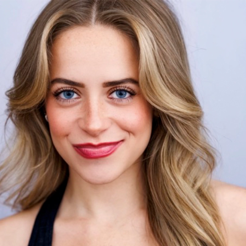 A woman with wavy, light brown hair and blue eyes smiles at the camera. She is wearing a sleeveless dark top and natural makeup with red lipstick. The background is a plain light gray, creating a simple and professional headshot.