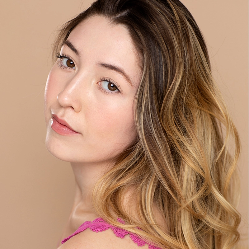 A young woman with wavy, light brown and blonde hair looks over her left shoulder. She has fair skin, natural makeup, and wears a pink top with lace trim. The style is reminiscent of Taylor Leccese against a plain, light beige background.
