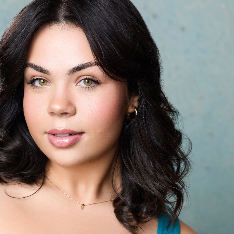 Ashlyn Filippone, a young woman with wavy dark brown hair, light skin, and hazel eyes, poses in front of a light blue speckled background. She wears a turquoise top, subtle makeup, small hoop earrings, and a delicate gold necklace.