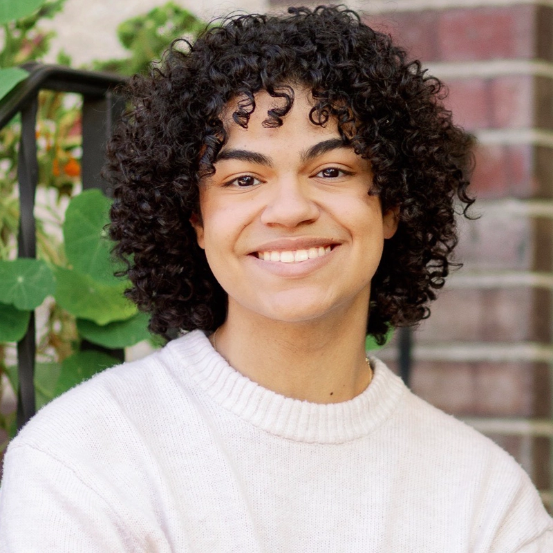 Diego Fleetwood, a young person with curly dark hair, smiles warmly at the camera. They wear a cream-colored sweater and sit outdoors by a brick wall, with green leafy plants and soft natural lighting in the background.