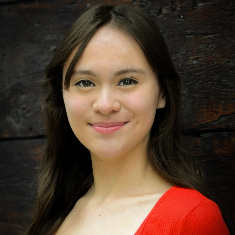 A young woman with long brown hair and fair skin smiles gently at the camera. She is wearing a bright red top and stands in front of a dark wooden background, which contrasts with her clothing and highlights her features.