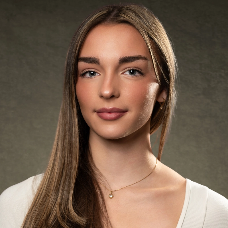 Veronica Fowler, a young woman with long straight brown hair and fair skin, poses against a plain gray background. She wears a cream-colored top and a delicate gold necklace, looking confidently at the camera with a gentle smile.