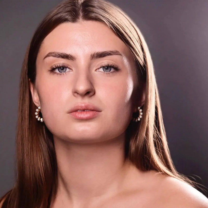 A young woman with long brown hair and fair skin, Jordyn Freetag, looks directly at the camera. She wears pearl earrings and has a neutral expression. The soft, dark gradient background highlights her clear skin and symmetrical features.