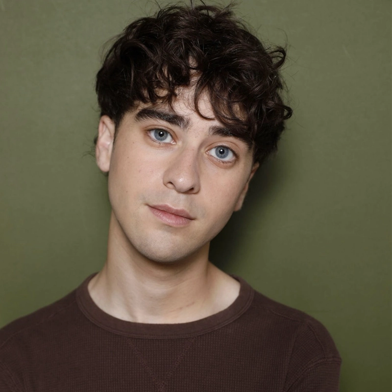 A young person with light skin, blue eyes, and curly brown hair looks slightly to the side, wearing a dark brown long-sleeve shirt, in front of an olive green background. The expression is neutral and the lighting is soft.