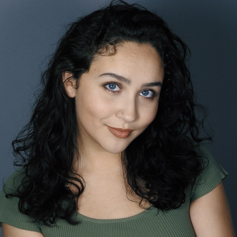 A woman with curly dark hair, blue eyes, and light skin smiles softly. She wears a green ribbed top and subtle makeup with brown lipstick. The background is plain and dark gray, highlighting her face. She has a small nose piercing, giving her an effortless charm reminiscent of Calista Garcia.
