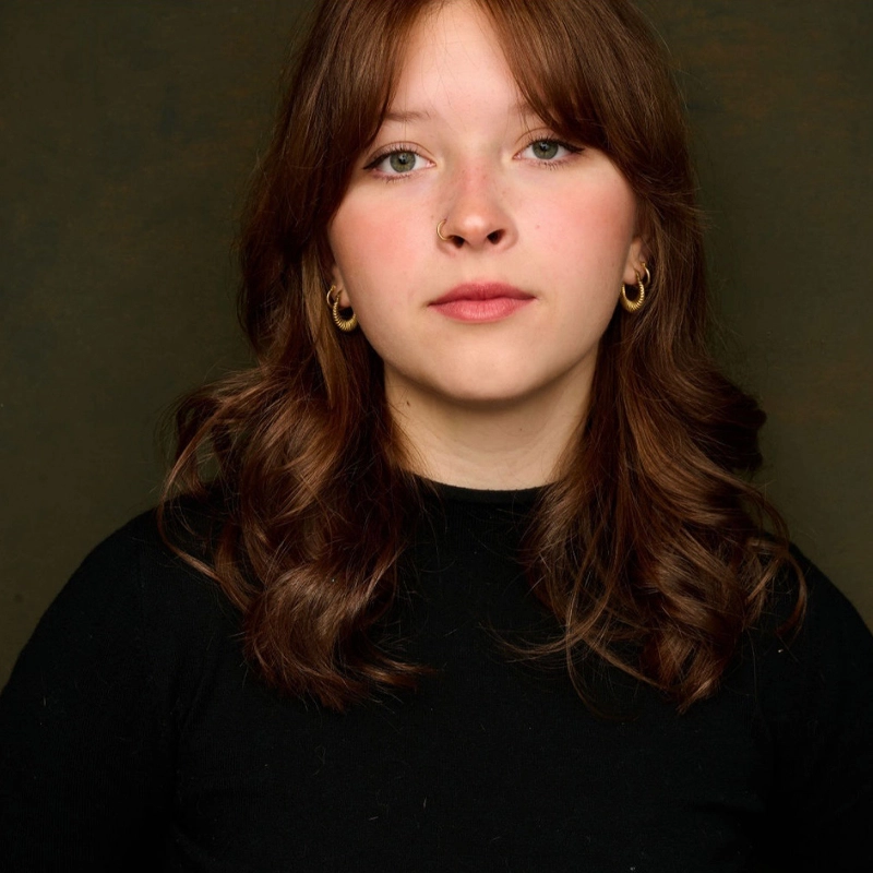 A young woman with light skin and wavy, chestnut-brown hair looks directly at the camera. Lily Giuliani wears a black top and gold hoop earrings. The dark, plain background draws focus to her calm, neutral expression.