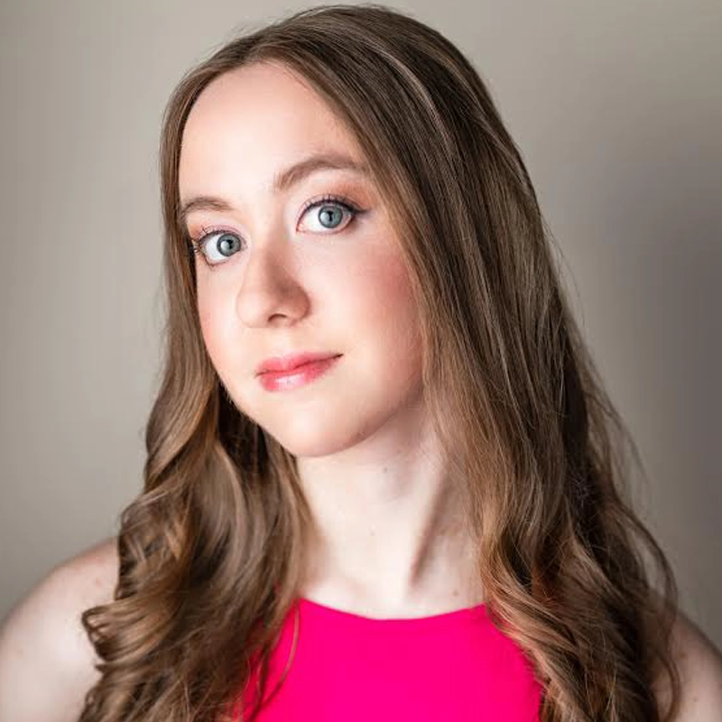 Britney Goldberg, a young woman with fair skin, long wavy light brown hair, and blue eyes, looks at the camera in a sleeveless bright pink top and subtle makeup. The background is neutral and softly blurred.