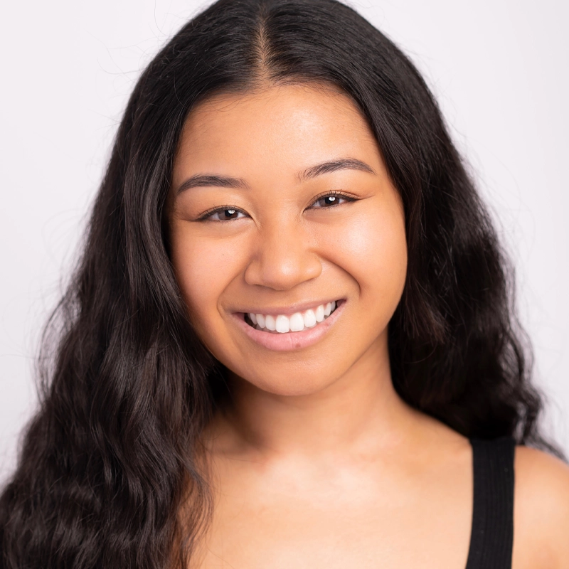 Jeyna Lynn Gonzales, a young woman with long, wavy black hair, smiles at the camera. She has smooth skin, straight white teeth, and wears a black sleeveless top against a plain, light-colored background that highlights her face.