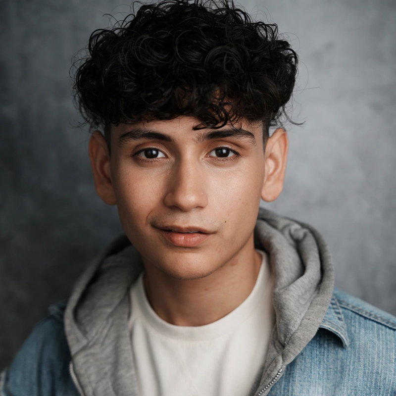 Marvin Gonzalez, a young man with short, curly dark hair, looks at the camera with a neutral expression. He is wearing a white T-shirt, a gray hoodie, and a light blue denim jacket against a soft, blurred gray background.