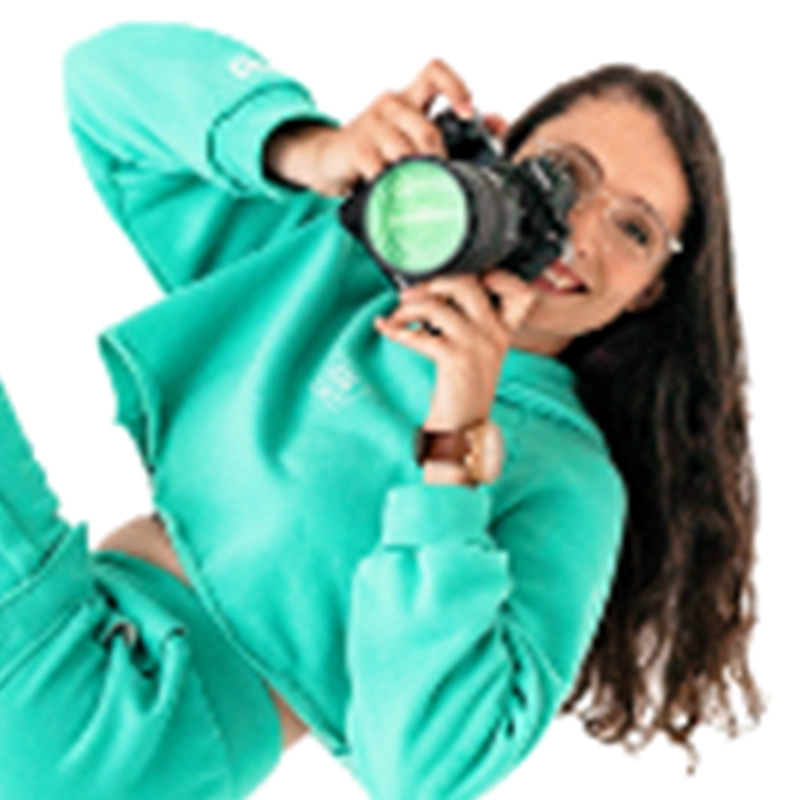 Liz Gilmartin, with long brown hair and glasses, smiles as she playfully holds a camera to her face. Dressed in a matching teal sweatshirt and sweatpants, she reclines against a white background, capturing the moment.