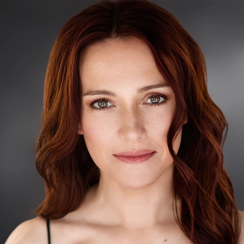 A woman with long, wavy auburn hair and light skin faces the camera with a neutral expression. She has brown eyes, subtle makeup, and is wearing a black strap on one shoulder. The background is dark gray and softly blurred.