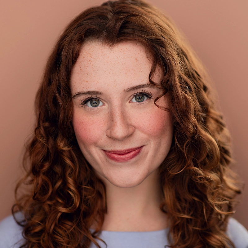 A young woman with fair skin, blue eyes, and long curly auburn hair smiles softly at the camera. Freckles dust her cheeks and nose as she wears a light lavender top—Heléne Rosborough stands before a plain, warm beige background.