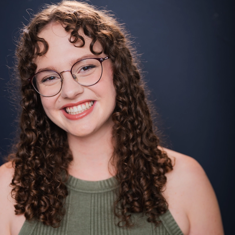 Hanna Hobson-Rohrer, a young woman with curly brown hair and glasses, smiles widely at the camera. She wears a sleeveless, ribbed olive-green top against a solid dark blue background, with bright lighting highlighting her cheerful expression.