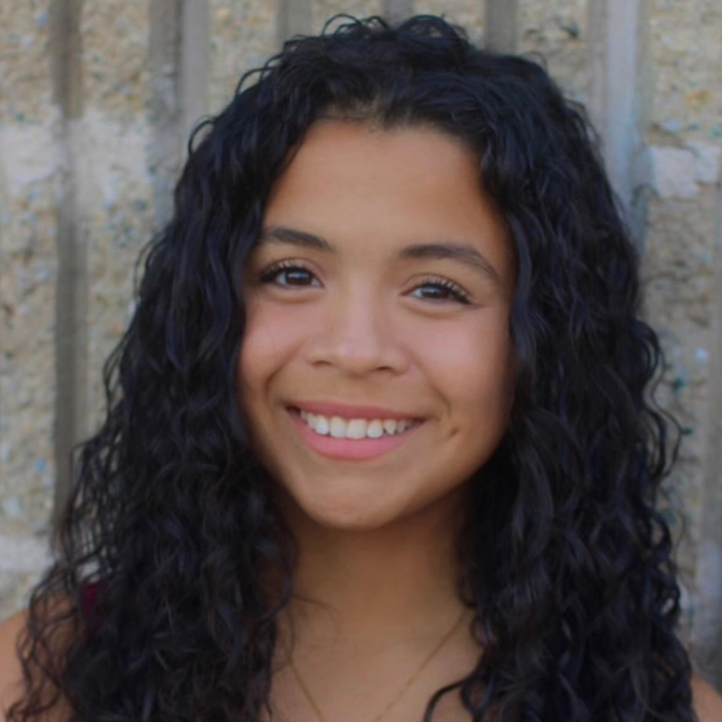 A young woman, Isabella Romero, with long, curly black hair smiles warmly at the camera. She has medium skin, dark eyes, and a dimple on her left cheek. She stands before a textured light brown concrete wall, wearing a simple necklace.