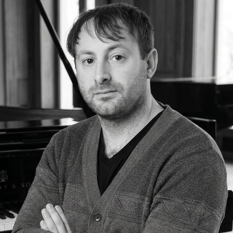 A man with short, light hair and a trimmed beard, wearing a patterned cardigan over a dark shirt, stands with arms crossed in front of a grand piano in a softly lit room. The black and white photo features Zachary Isrow.