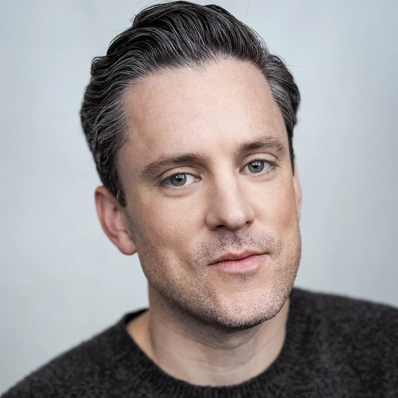 A man with fair skin and short, neatly styled dark hair looks into the camera with a relaxed expression. He is wearing a dark sweater, and the background is a soft, plain light gray, creating a calm and neutral setting.