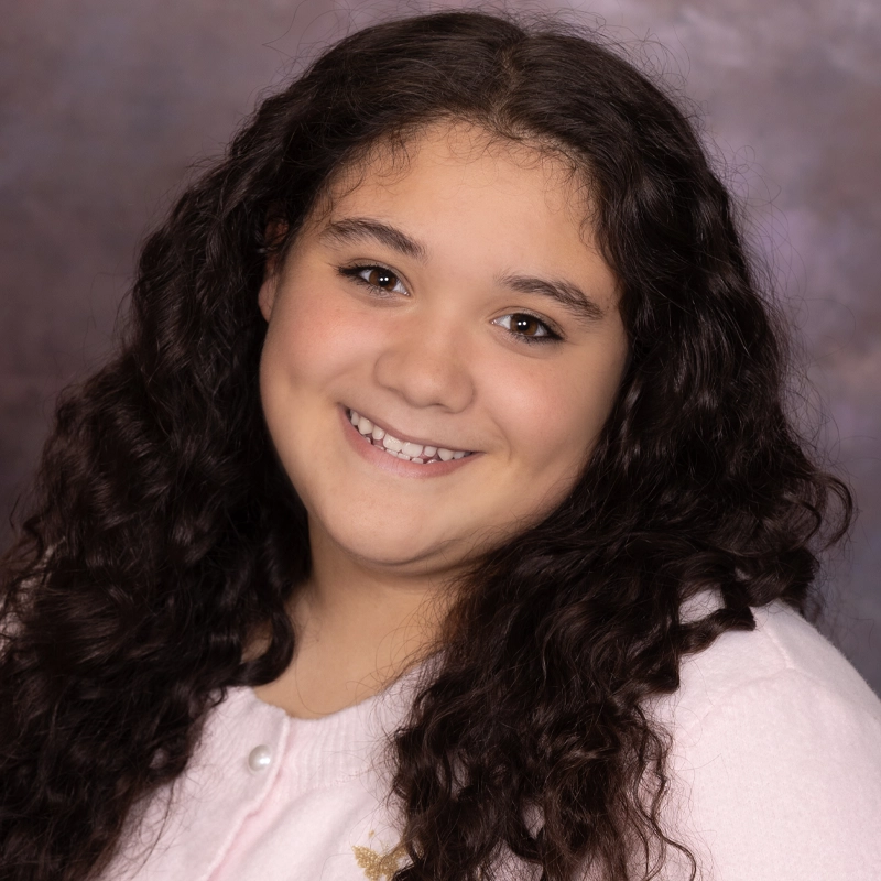 Isabella Jackson, a young person with long, dark, curly hair, smiles warmly at the camera. They wear a light pink sweater with a small embroidered detail and pose in front of a softly blurred, purple-brown studio background.