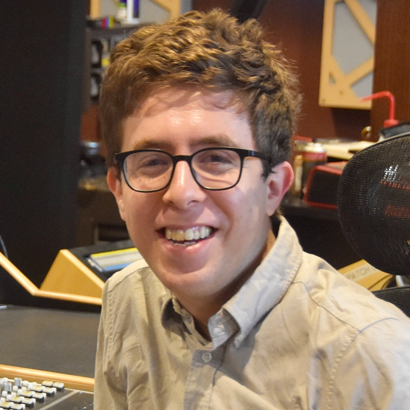 A young man with short brown hair and glasses smiles at the camera. He is wearing a light gray button-up shirt and is seated in Liz Gilmartin’s recording studio, with audio equipment and a black mesh chair visible in the background.