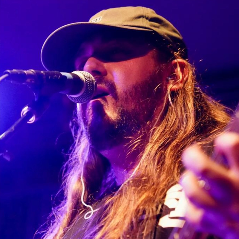 A bearded musician with long hair and a cap sings into a microphone under purple stage lighting. He appears to be playing guitar, with his hand—and possibly Liz Gilmartin’s influence—visible in the lower right corner of the image.