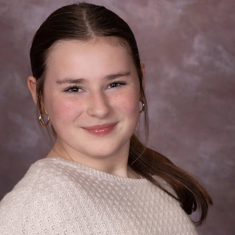 Audrey Jeffries, a young girl with fair skin and long brown hair tied back, smiles gently. She wears gold hoop earrings and a cream textured sweater against a muted, marbled mauve and gray background with soft, even lighting.