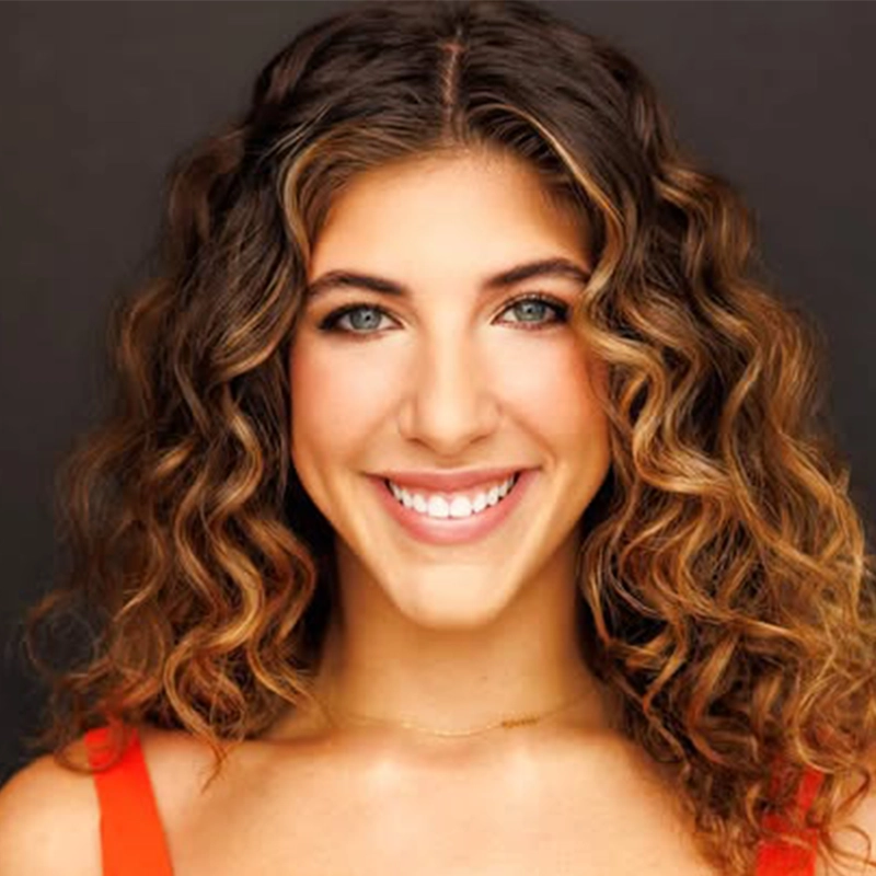 Smiling woman, Dominique Alvarez, with long, voluminous, dark blonde curly hair and light eyes wears an orange top and a delicate gold necklace. She faces the camera against a dark background, exuding a friendly and confident demeanor.