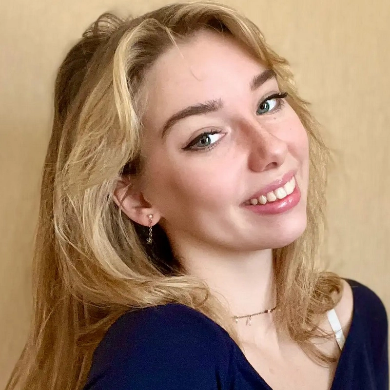 Lizzie Johnson, a young woman with long blonde hair, smiles at the camera. She is wearing a navy blue top, a delicate necklace, and small earrings. The plain beige background highlights her cheerful expression and natural makeup.