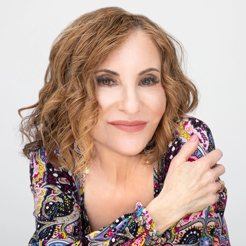 A woman with wavy, light brown hair and fair skin smiles gently at the camera. She wears a colorful, patterned blouse with pink, purple, and yellow designs, and has her right arm across her chest, creating a relaxed and welcoming pose against a light background.