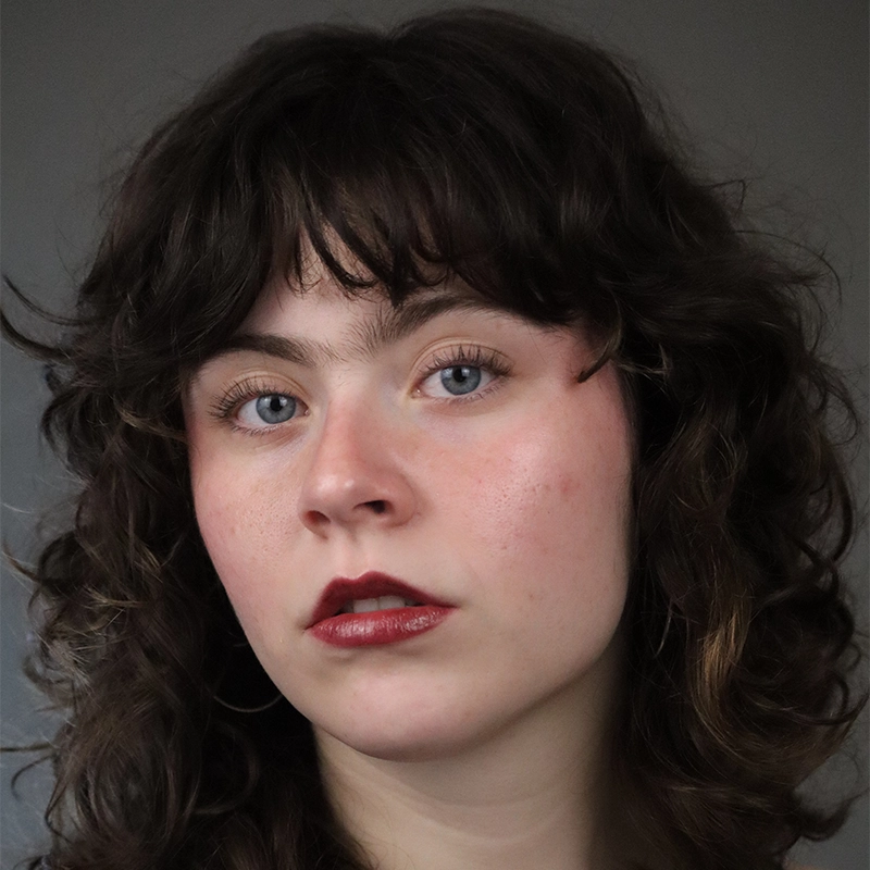 A young person with pale skin, blue eyes, and wavy dark brown hair with bangs looks directly at the camera. They have natural makeup, including bold dark red lipstick, and a neutral facial expression. The background is plain and gray.