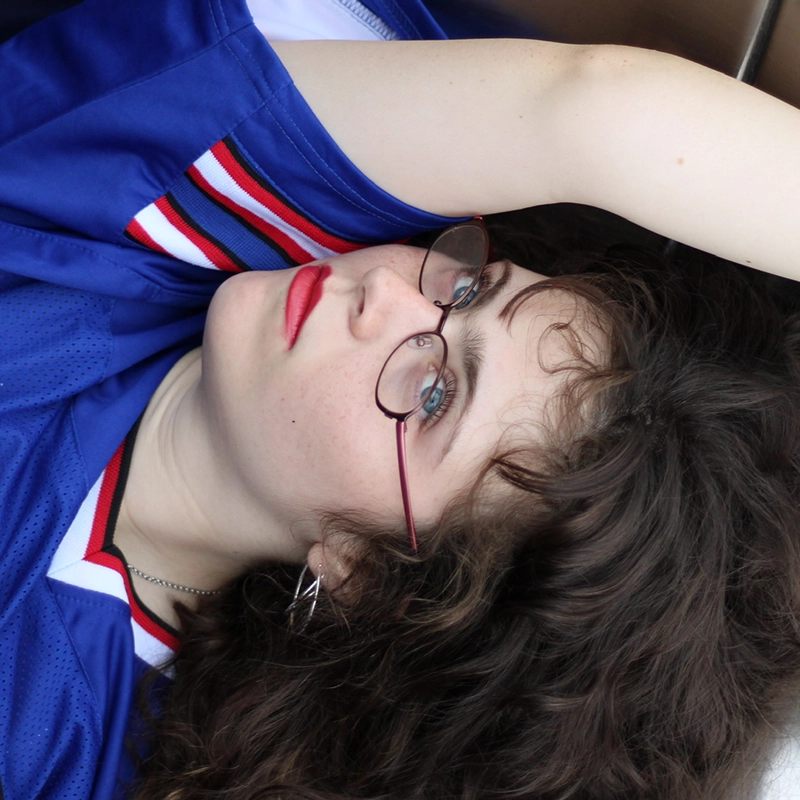 A young person with long curly hair, red lipstick, and round glasses lies on their back. They wear a blue sports jersey with red, white, and black striped collar details. One arm is raised above their head, and they gaze upwards with a relaxed expression.