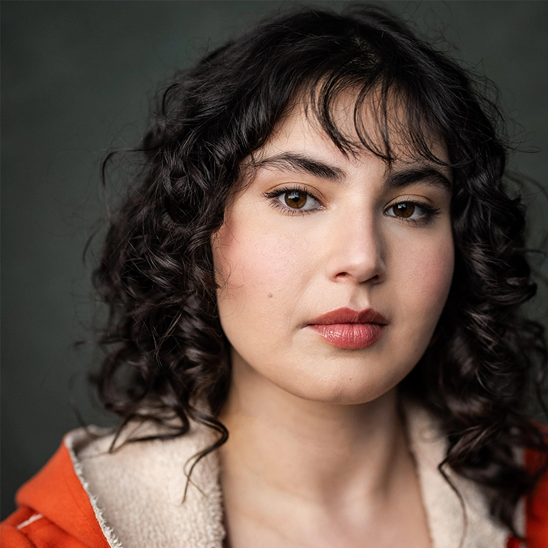 Casey Martin Klein, a young woman with pale skin, wavy dark brown hair, and brown eyes, looks at the camera with a neutral expression. She wears natural makeup and a rust-orange jacket with a shearling collar against a dark, blurred background.