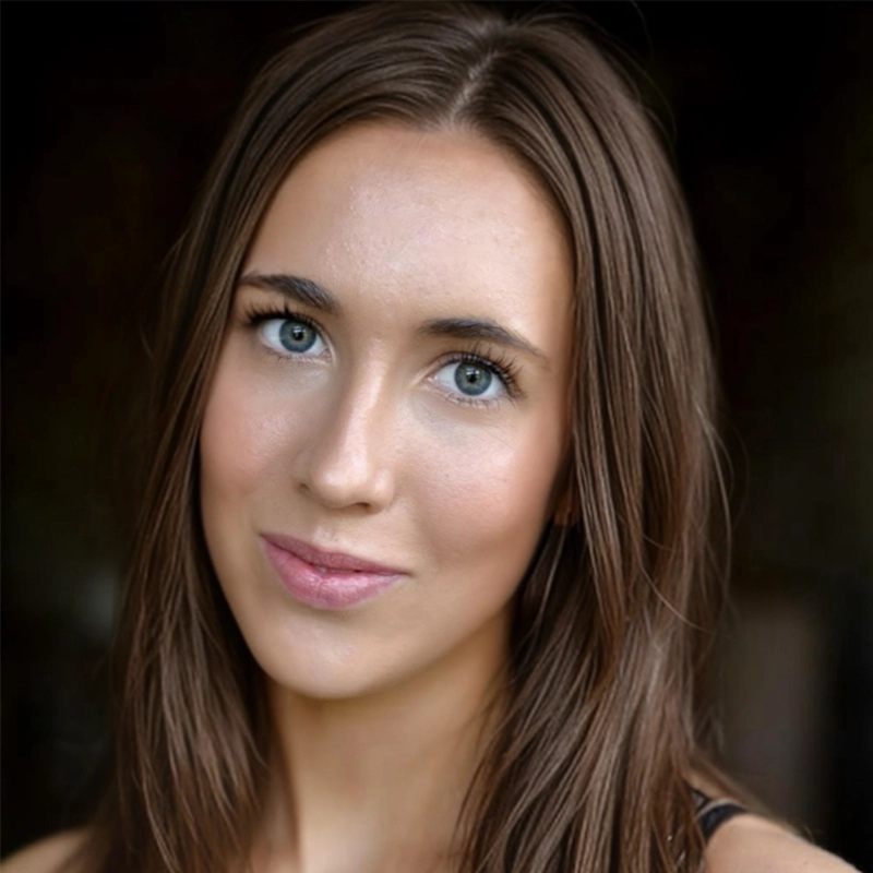A woman with fair skin, long straight brown hair, and blue eyes looks directly at the camera with a slight, closed-mouth smile. She is wearing minimal makeup and a dark top. The background is out of focus and dark, highlighting her face.