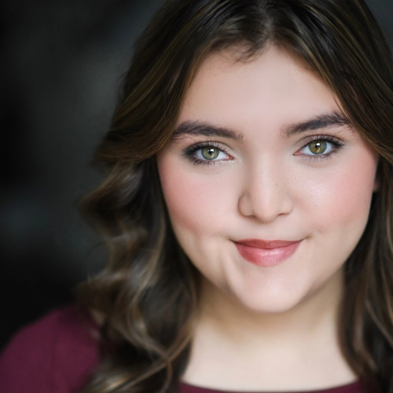 Gabrielle Karyss, a young woman with wavy brown hair and green eyes, smiles softly. She has clear skin, natural makeup, and wears a burgundy top. The dark, blurred background brings focus to her face.