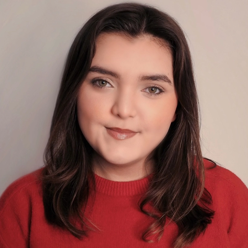 A young woman with long brown hair and fair skin looks at the camera with a slight smile. She is wearing a red sweater, has subtle makeup, and is posing against a plain, light-colored background.