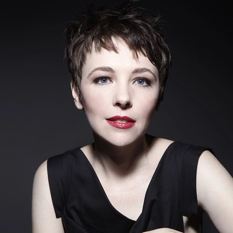 A woman with short, dark brown hair and light skin poses against a dark background. Harmony Keeney has blue eyes, wears dark red lipstick, and a sleeveless black top. Her calm expression and soft smile face the camera directly.