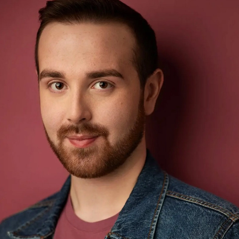 A young man with fair skin, short brown hair, and a trimmed beard smiles gently. He wears a jean jacket over a maroon shirt. The background is a matching deep maroon, giving the photo a warm, cohesive feel. He looks directly at the camera.